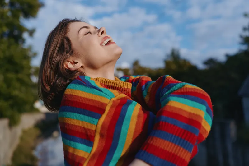 woman smile rainbow sweater outdoors portrait joyful Young woman in a rainbow striped sweater laughs with eyes closed looking up outdoors, candid portrait showing joy, colorful fashion, relaxed