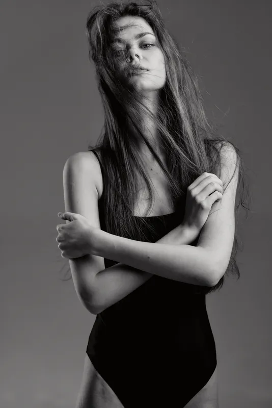 Black and white portrait of a young woman with long hair wearing a black bodysuit, expressing calm and confidence in studio lighting.