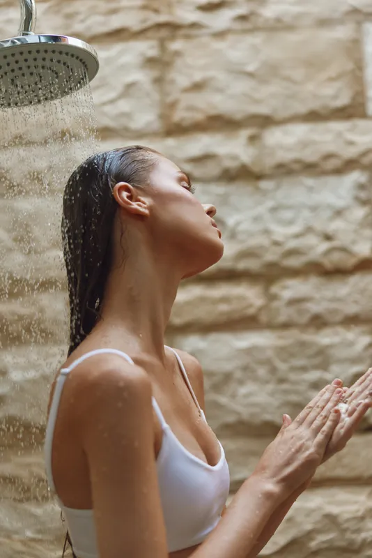 woman showering outdoors under rain spray, wet hair, white swimsuit top, natural stone wall background, refreshing moment, summer bath