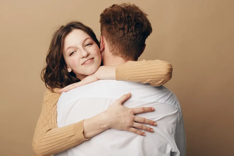 Young woman in beige sweater hugs man in white shirt with closed eyes and gentle smile on beige background. Concept of love, affection, relationship, and emotional connection.
