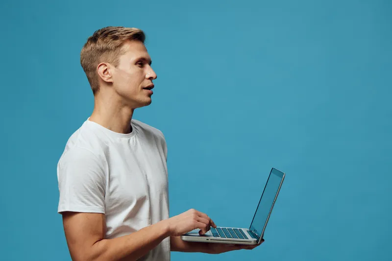 serious young caucasian man working laptop hold stand side view blue background casual white t-shirt indoor digital nomad concept