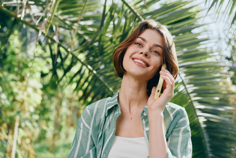 Portrait of a cheerful woman talking on a mobile phone amid lush greenery, sunlight, and a relaxed casual vibe.