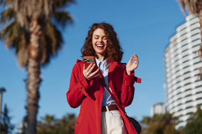 Young woman in a red trench coat laughs while looking at her smartphone outdoors under a bright blue sky, with palm trees and a modern building in the background, a candid cheerful moment.
