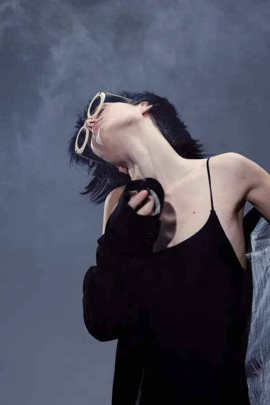 Stylish woman with short hair wearing sunglasses and black outfit posing against smoky background.