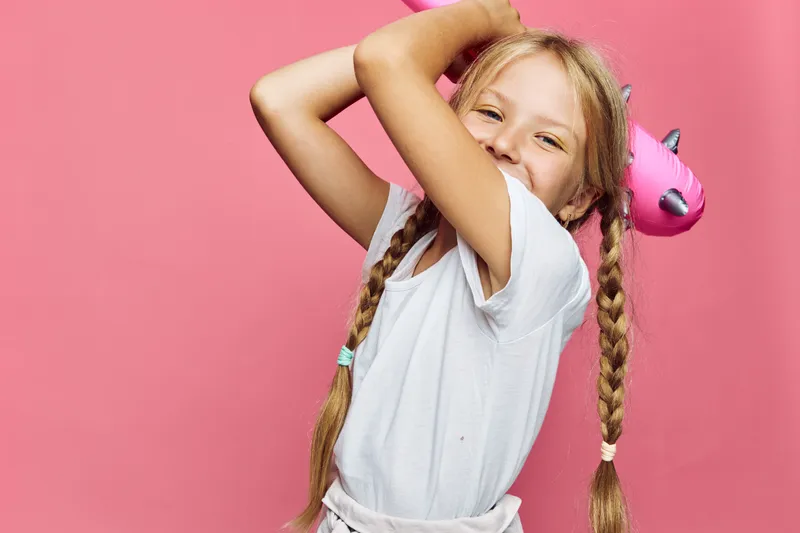 Happy young girl with braided hair smiling and holding a pink inflatable unicorn float on a pink background. Concept for childhood, summer, and fun activities.