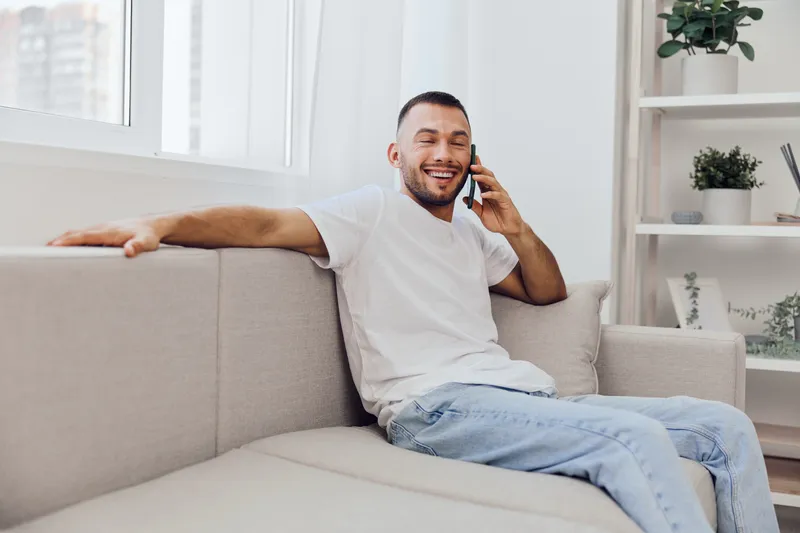 Relaxed man enjoying a phone call on a cozy couch, embodying the essence of soft masculinity and emotional vulnerability in a serene home environment.