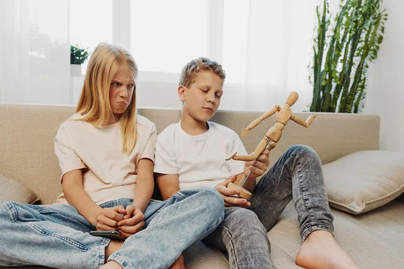 Annoyed girl and calm boy sitting on couch with wooden mannequin, expressing different emotions in cozy living room setting.