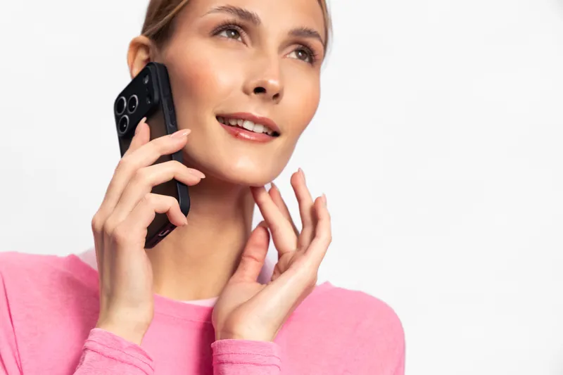 Young woman in pink sweater talking on smartphone with smile, looking away. Bright studio lighting highlights her natural skin and casual positive mood, concept for communication and technology.