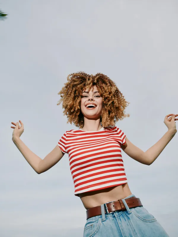 Joyful woman with curly hair wearing a red striped crop top and denim jeans, arms raised, smiling under a clear sky, capturing casual fashion and sunny energy.