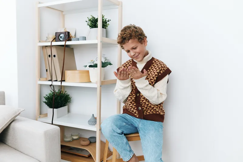 Young boy sits on a wooden chair, knitting with concentration in a bright, cozy living space. Soft light and tidy shelves create a warm, homey atmosphere.