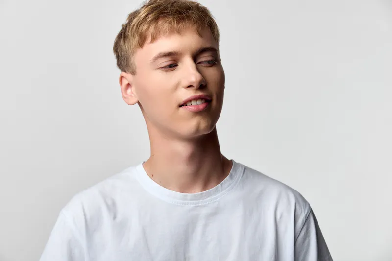 Bright studio shot of a young man in a white shirt with a casual, relaxed expression, looking slightly to the side, clean neutral background, simple lighting, suitable for lifestyle, fashion, or