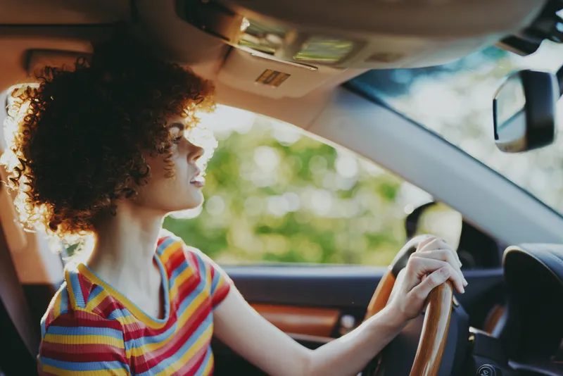A woman driver in a striped shirt sits behind the wheel in a sunlit car interior, capturing a casual, retro lifestyle mood with warm film color and nostalgic flair.