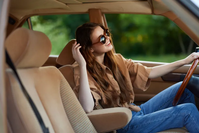 Young woman enjoying a summer drive in a stylish vintage car, featuring a warm color palette, smiling, with sunglasses and casual clothing.