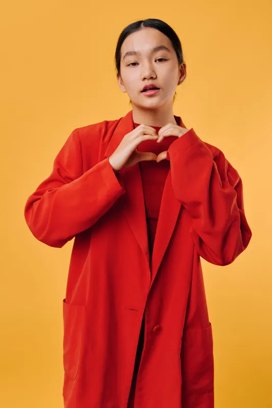 Woman in red jacket standing on yellow background making heart shape with hands, casual portrait and positive emotion with simple vibrant style and gesture for love.