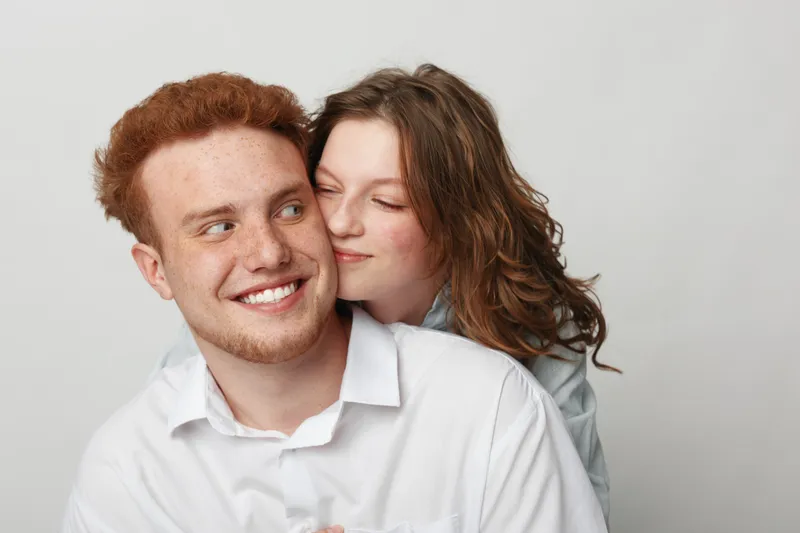 Smiling young couple in white shirts embraces tenderly against a plain background. The woman gently kisses the man’s cheek, expressing love and affection in a warm moment.