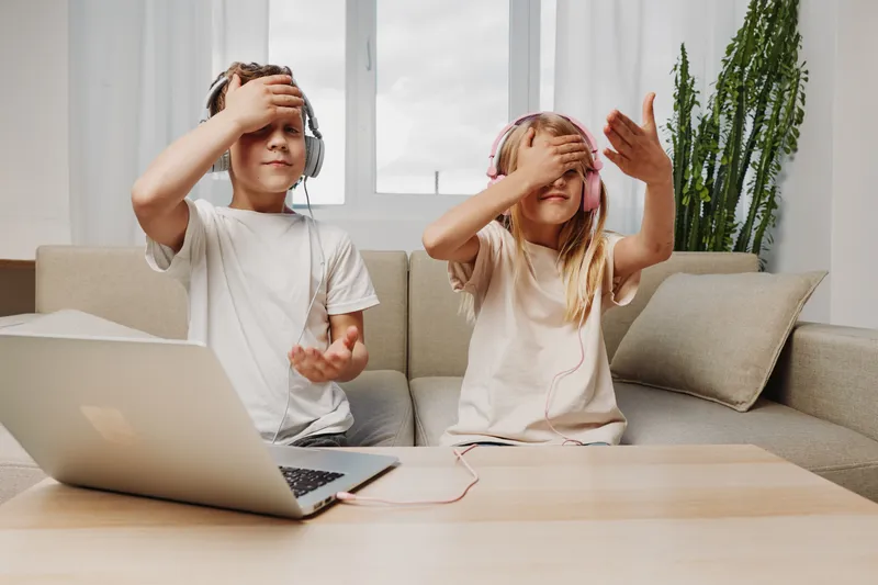 Two children sitting on sofa with headphones, playing a game with eyes covered by hand. Modern living room, laptop on table, concept of fun, family, and digital entertainment.