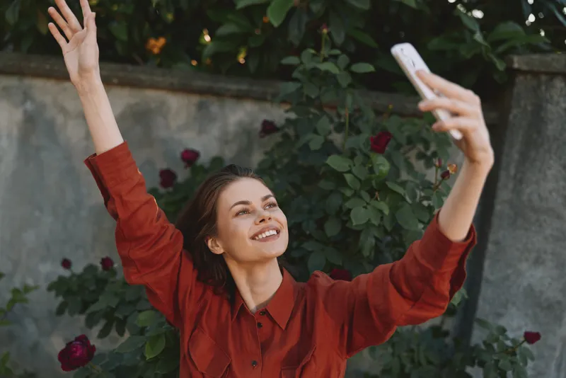 Cheerful woman outdoors, taking a selfie with a bright smile, casual red top, sunlit garden background, lush greenery, relaxed pose, joyful lifestyle moment, vibrant ambiance, candid