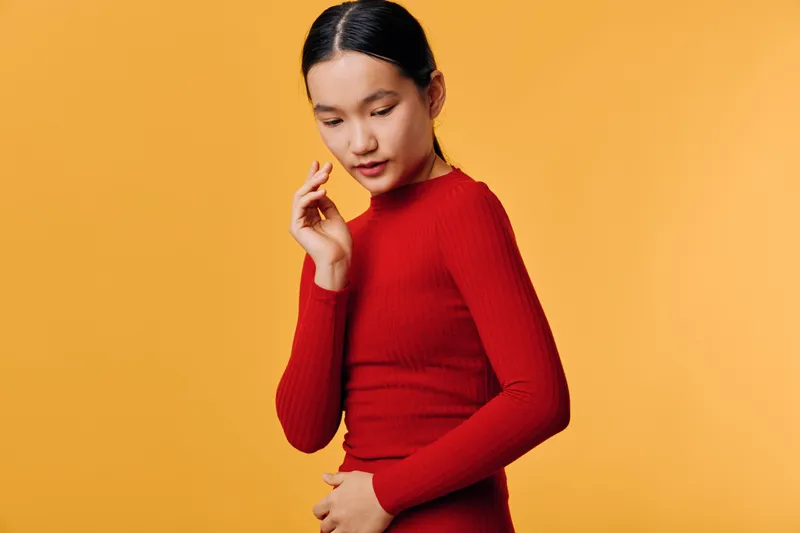 Young woman in red sweater standing against yellow background with calm expression. Female model with dark hair in neat hairstyle posing thoughtfully for portrait photography.
