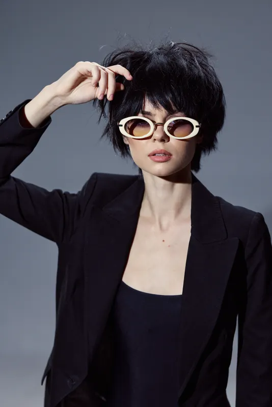 Stylish woman with short black hair wearing white oval sunglasses and a black blazer posing against gray background