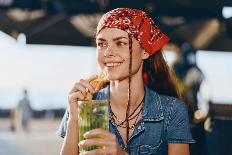 Lifestyle woman in modern boho-western denim street style sipping a refreshing drink, natural smile, casual summer vibe, warm film color tones captured outdoors.
