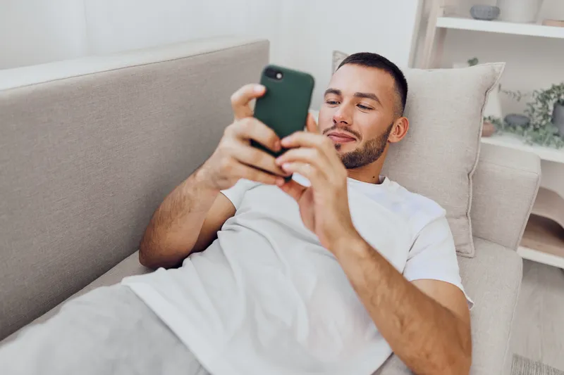 Relaxed man using smartphone on a cozy couch, engaged in social media or browsing the internet, enjoying leisure time in a modern living space.