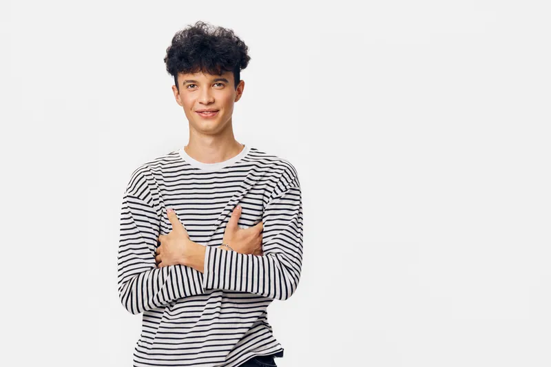 young man in striped shirt smiling with crossed arms on white background, casual style, positive expression, studio portrait with soft lighting and minimalistic setting.