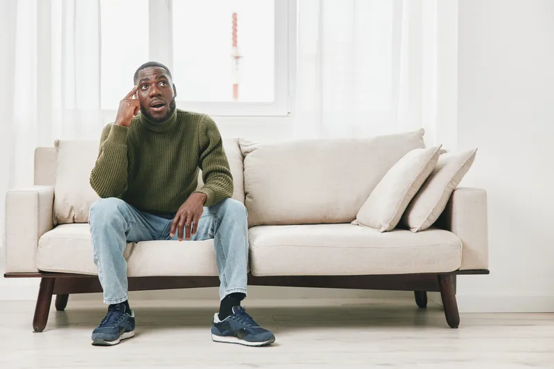 Concerned young man sitting on a sofa, expressing anxiety while talking on the phone in a cozy, bright living room setting with natural light