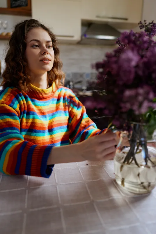 Woman indoors at a kitchen table, wearing a colorful striped sweater, relaxed mood, playful moment, flowers in a vase, soft natural light, homey atmosphere