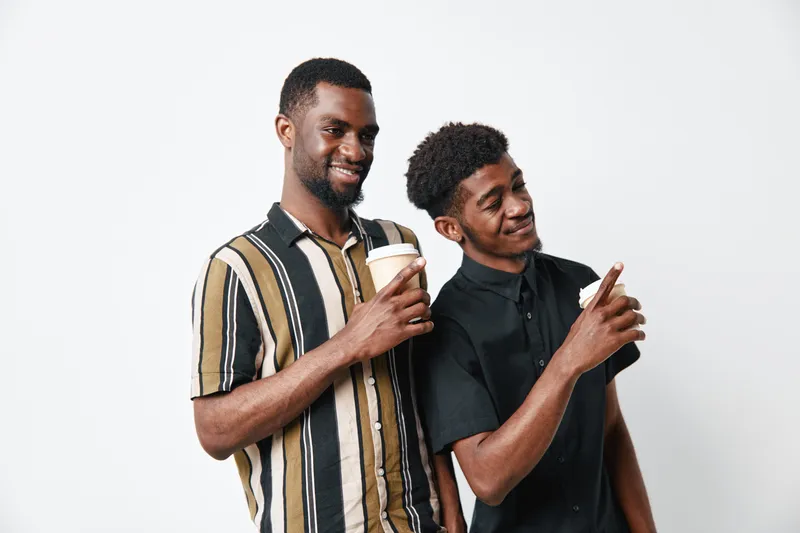 Two African American men with dark skin smiling and holding coffee cups, casual clothing, standing together on white background, friendship and relaxation concept.