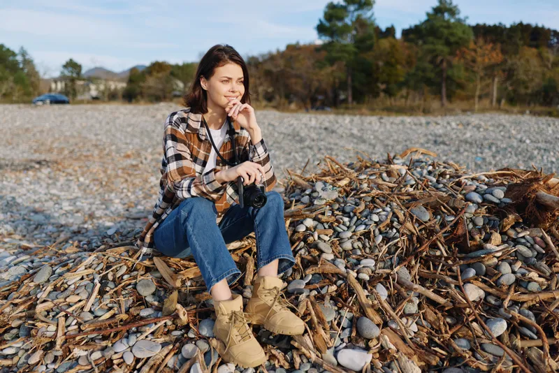 Woman outdoors on a pebbled beach, autumn vibes, plaid shirt and boots, sitting contemplatively among seaweed and stones along a calm shoreline, a lifestyle portrait in serene coastal scenery