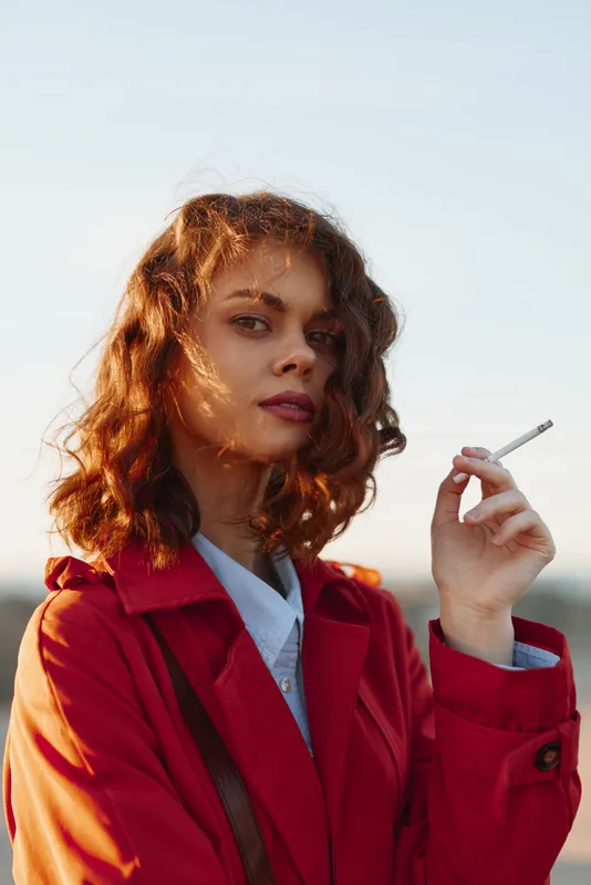 Young woman with curly brown hair stands outdoors in a red coat, holding a cigarette with a relaxed pose and confident gaze under clear daylight.