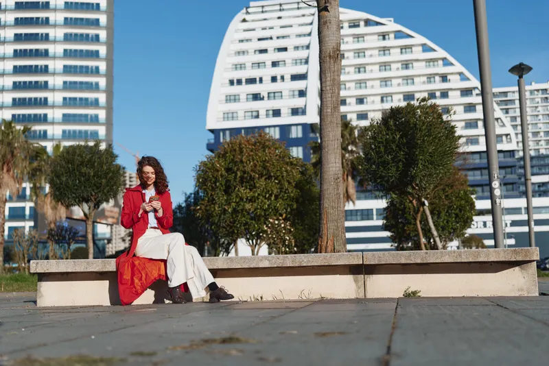 Young woman with curly hair sits on a concrete bench in an urban park wearing a red coat and a white dress, capturing a calm stylish moment amid tall modern buildings.