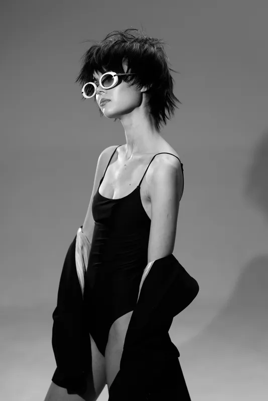 Black and white portrait of a stylish woman wearing sunglasses and a black dress with jacket, posing confidently indoors.