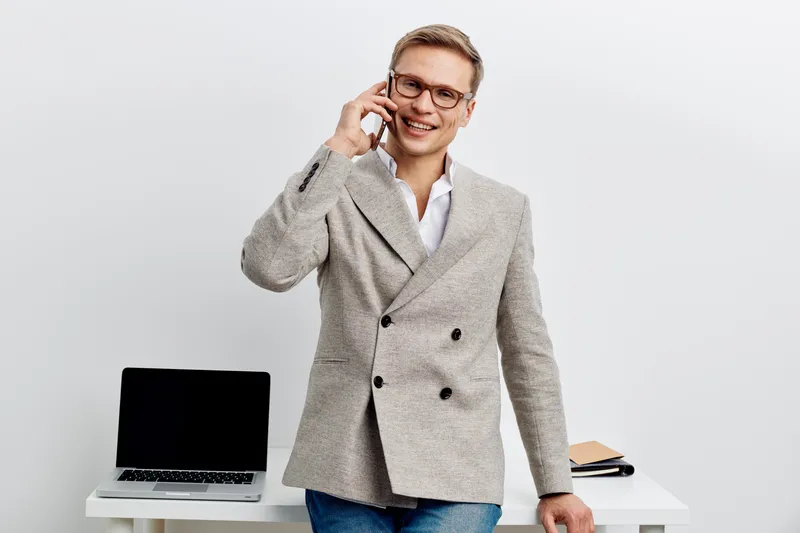 Smiling professional man in blazer talking on mobile phone in office interior with laptop and documents on desk, business concept, modern workplace, casual style, positive attitude, young adult, profe