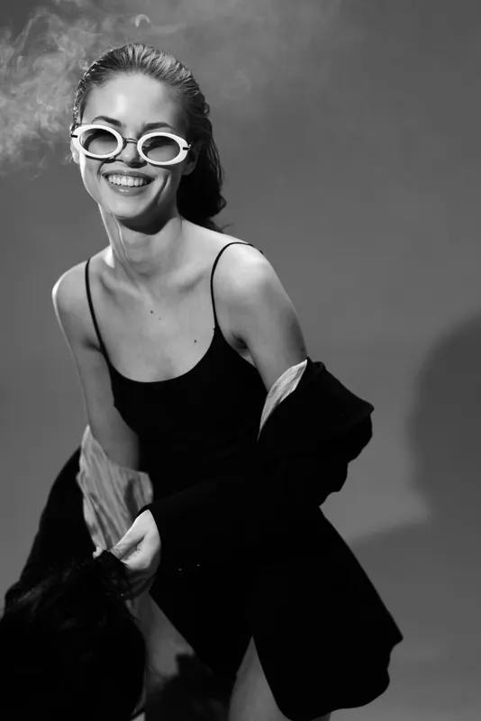 Smiling woman wearing sunglasses and stylish clothes in black and white portrait with joyful expression and relaxed pose.