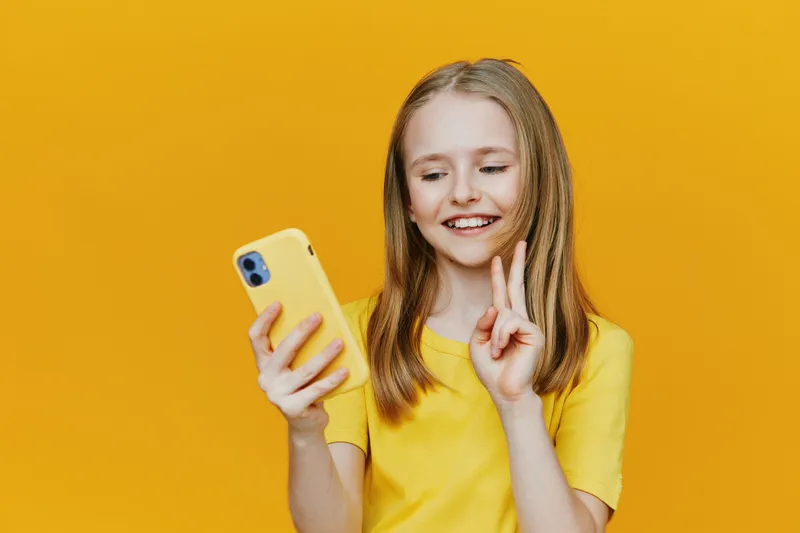 Cheerful young girl with long hair holding a phone, wearing a yellow t shirt, against a vibrant yellow background, expressing happiness and positivity in a seasonal setting