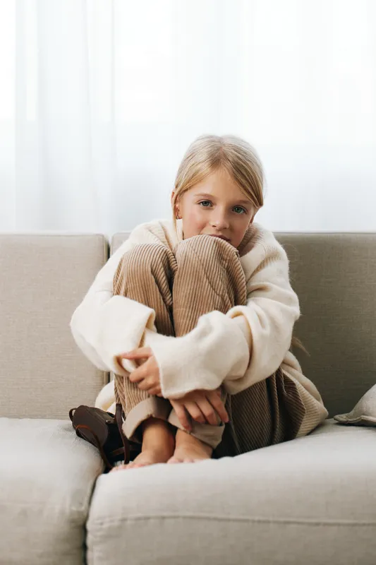 Authenticity and school lifestyle in a cozy home setting, a child sits on a sofa, knees hugged, wearing a sweater and knit pants, a quiet moment of everyday imagination.