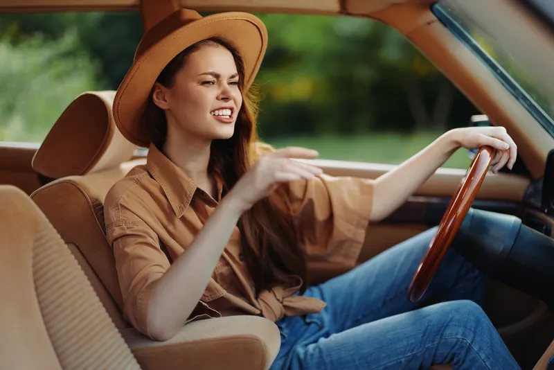 Young woman driving a vintage car, expressing joy and excitement in casual outfit with a hat, surrounded by nature's vibrant greenery.
