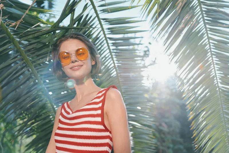 A cheerful woman wearing a red and white striped top and round sunglasses smiles under bright sun among tropical palm fronds, creating a lively outdoor vibe.