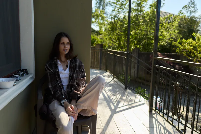 Casual woman outdoors, seated on balcony, plaid shirt and light trousers, relaxed pose, soft sunlight, greenery background, natural colors, everyday lifestyle moment
