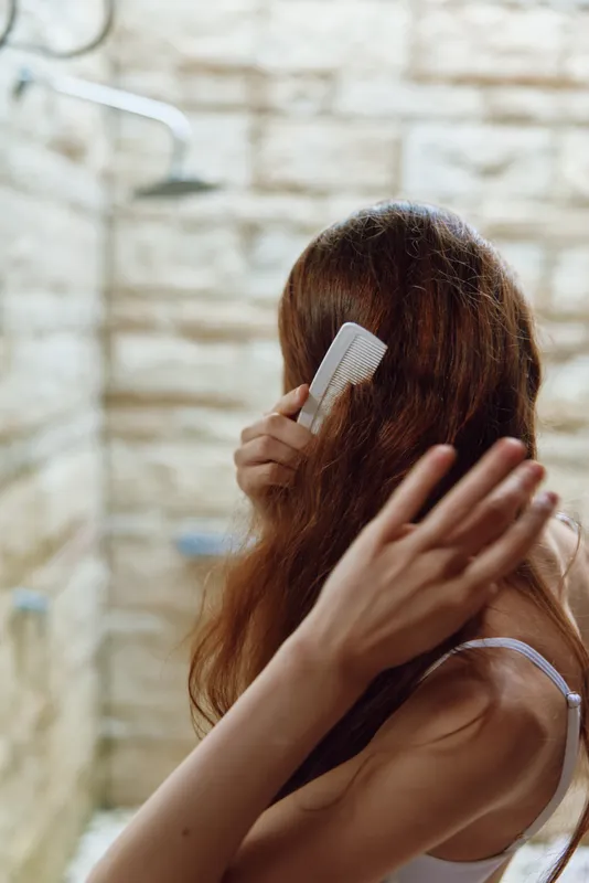 woman combing long brown hair with white comb in bathroom with beige stone wall, soft natural light, intimate daily routine moment