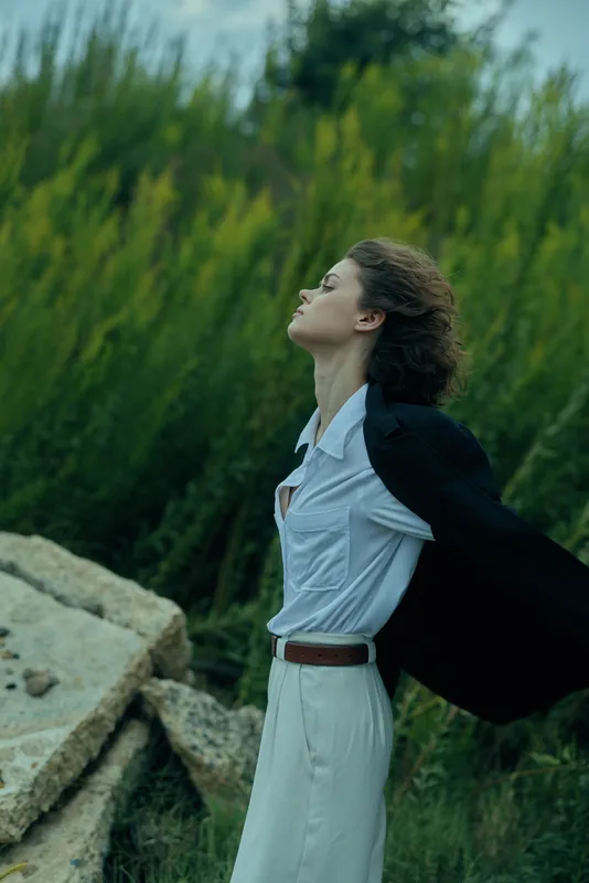 Woman outdoor dress cape wind nature in a tranquil moment, a white dress flows as the cape billows, she gazes upward, greenery and rocks frame the scene, expressing freedom and calm in open air.