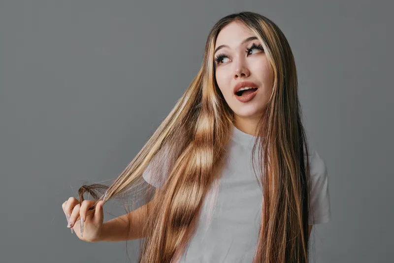 Young woman with long straight hair wearing light shirt posing with playful expression on grey background in studio. Beauty, fashion, hairstyle and lifestyle concept.
