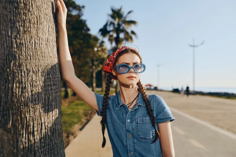 Lifestyle woman in modern boho-western denim street style wearing blue sunglasses and red bandana, posing outdoors with palm trees and film color tones.