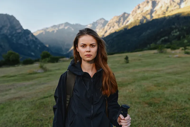 A focused woman stands in a mountain valley wearing a dark jacket and backpack, holding a camera, ready to explore the wild open air and capture dramatic nature.
