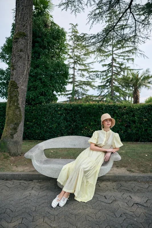 Elegant woman in a pale yellow dress and straw hat sits on a curved bench in a park, surrounded by trees, creating a calm outdoor portrait