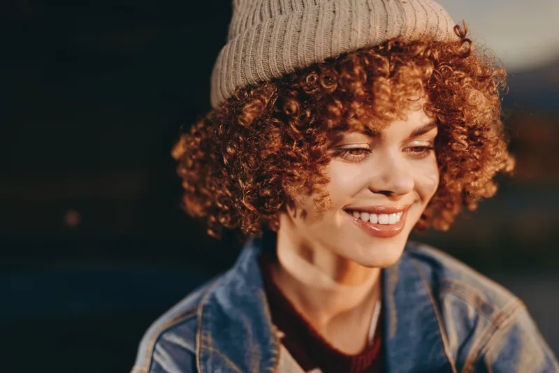 Woman with curly hair and smile wearing a knit hat and denim jacket, outdoor lifestyle portrait, warm golden evening light, soft shallow focus, cozy mood, friendly face