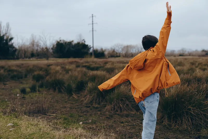 Woman outdoors in an orange jacket, walking through a grassy field, jeans visible, arms raised in windy air, cloudy sky, lifestyle moment, natural colors, peaceful mood, for wellbeing