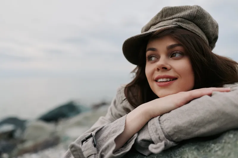 Smiling young woman wearing a brown cap rests her arms on a stone outdoors, looking to the side with a peaceful expression. Cloudy sky and blurred nature background create calm mood.