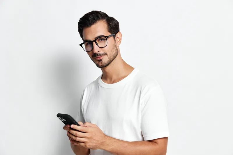 Handsome young man in white t-shirt and glasses standing with smartphone, smiling confidently at the camera against plain white background. Modern lifestyle concept.